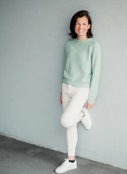 Eine Frau trägt einen mintgrünen Pullover und weiße Hosen, lächelt freundlich in die Kamera.