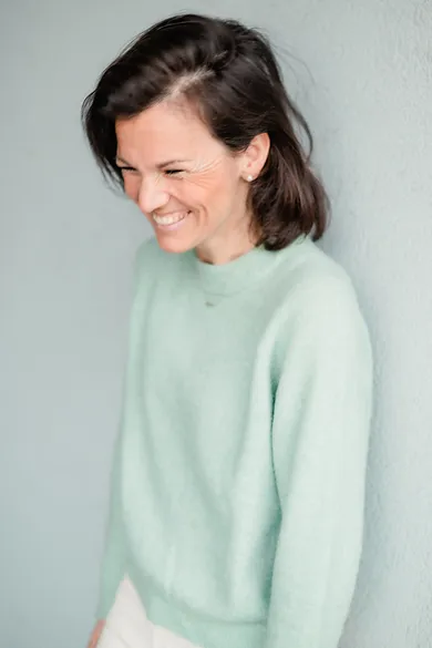 Eine Frau trägt einen mintgrünen Pullover und weiße Hosen, lächelt freundlich in die Kamera.